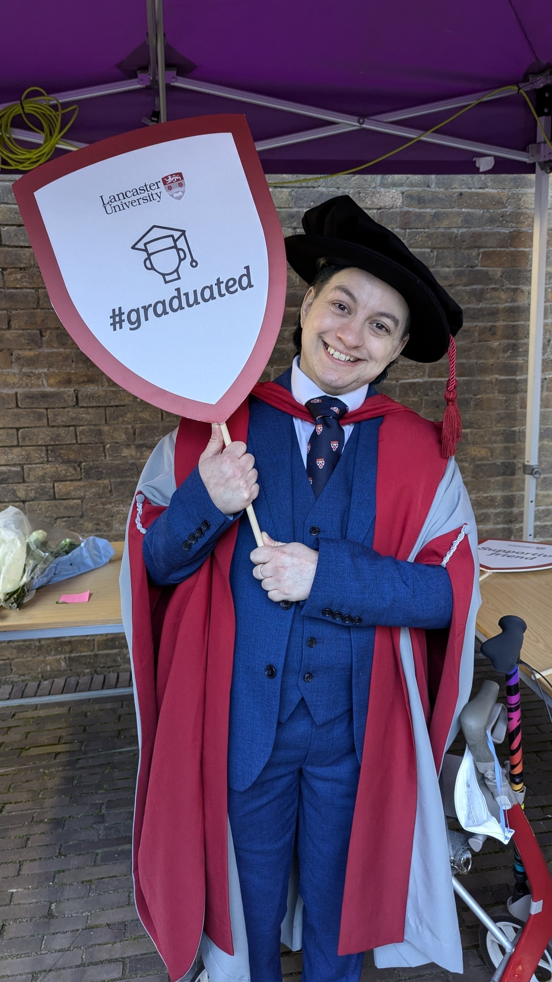 A person in academic regalia holds a sign with the Lancaster University logo and "#graduated" under a purple tent.