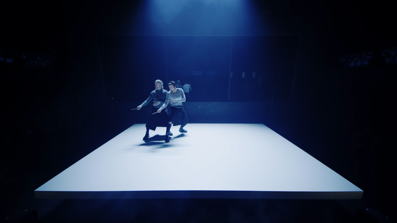 Cal Macaninch and David Tennant wearing black kilts and grey long-sleeved shirts, huddle in the far corner of a white stage while defensively wielding knives