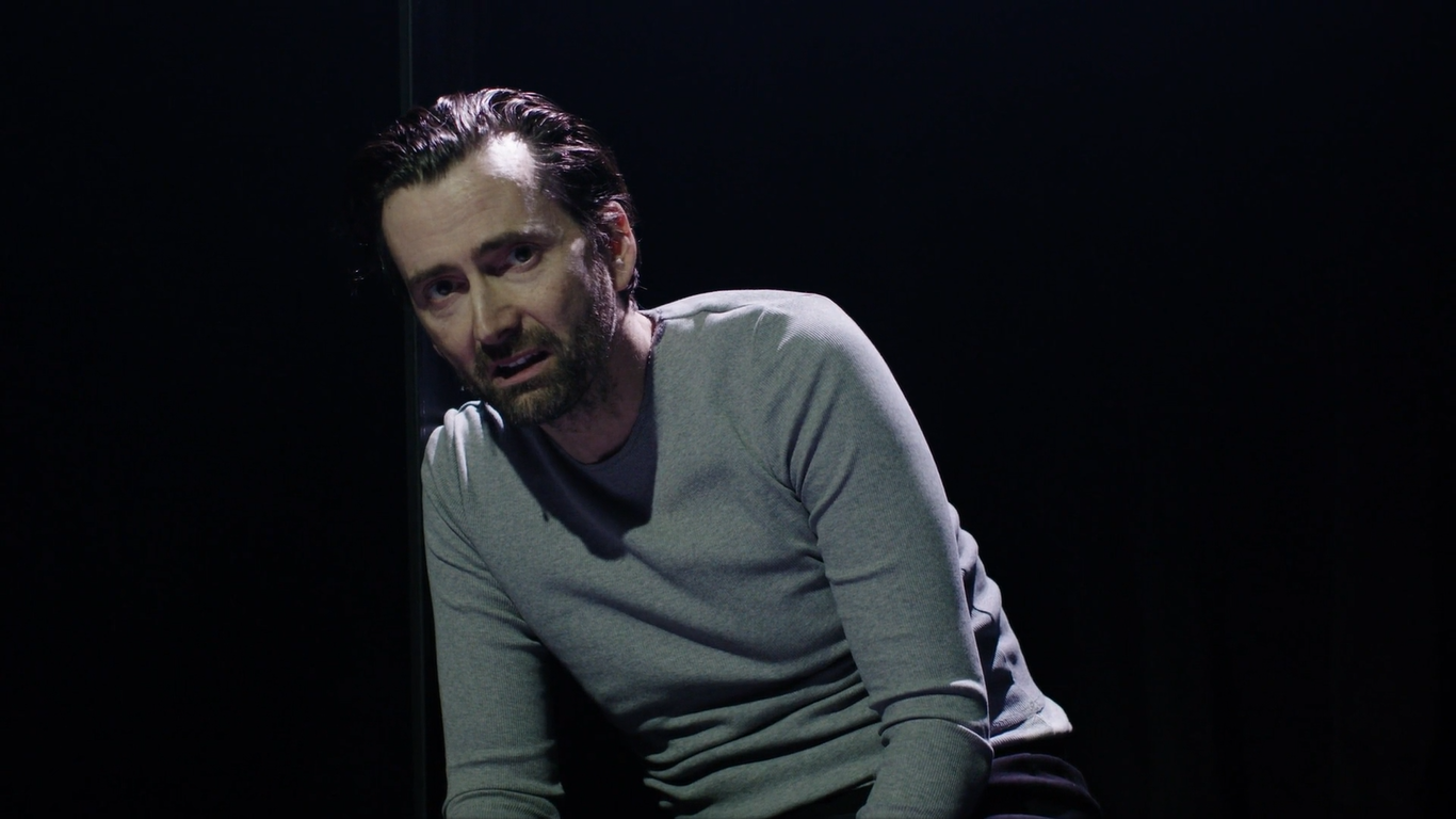 David Tennant as Macbeth looks wearily into the camera, elevated into the darkness above the stage