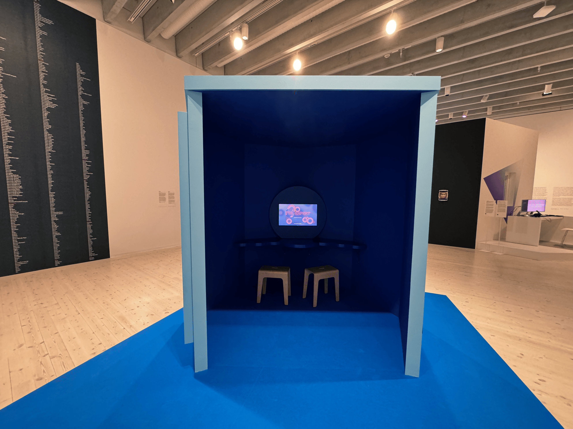 Installation: a booth with dark blue walls and two yellow stools, with a built-in desk and screen